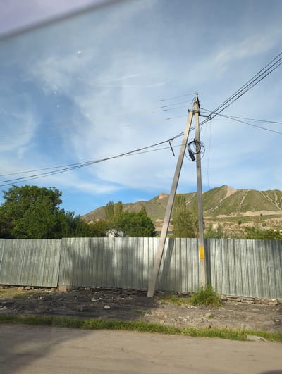 The scene is in Beş-Küŋgöy, Kyrgyzstan. A tilted view shows a weathered utility pole with numerous wires extending to the left and above, set against a blue sky with some clouds. To the right, a corrugated metal fence lines the edge of a paved road with a strip of grass. A rocky embankment with sparse vegetation separates the fence from a mountain visible in the background. Trees are interspersed between the mountain and the fence. A yellow warning sign is affixed to the utility pole near the fence. The perspective is taken from within a vehicle, as suggested by a partial view of the vehicle's interior in the lower left corner.