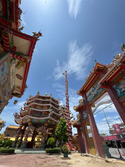The image depicts a brightly colored temple complex with ornate architecture against a clear blue sky with wispy clouds. The foreground features a red tiled courtyard leading up to the temple structures. To the left, a section of a richly decorated roof with dragon motifs and intricate paintings is visible. In the center, a towering, multi-tiered pagoda-like structure dominates the scene, supported by elaborate pillars adorned with dragons and detailed carvings. To the right, a large, red ceremonial gateway with a golden gate is prominent, featuring murals of tigers and other decorative elements. A tall, red, pole-like structure with a flag or streamer at the top stands to the left of the gateway. The background reveals more temple buildings, some with golden roofs, and hints of a street with buildings, traffic cones, and signage, suggesting an urban environment. The area is identified as Ang Sila, Thailand. There are no visible people or specific actions taking place.