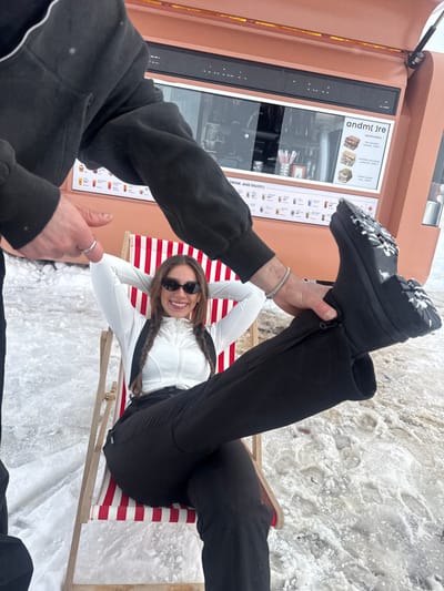 The image depicts an outdoor winter scene in a recreational area within Yeghipatrush, Armenia. A female subject, appearing to be a young adult, is seated in a red and white striped wooden deck chair. She is wearing dark oval sunglasses, a white long-sleeved zipped top, black ski pants with suspenders, and black, thick-soled snow boots. Her dark hair is styled in two braids, and she is smiling broadly, facing the camera with her hands clasped behind her head. Her left leg is extended upwards.

A second individual, partially visible from the forearms and hands, is positioned to the left of the frame, supporting the female subject's extended left leg. This individual's hands are grasping the ankle area of the female's left boot. The visible hands wear a silver ring on the fourth finger of the left hand and a silver bracelet on the right wrist. The individual wears a dark long-sleeved top, possibly a hooded sweatshirt.

In the background, an orange-brown mobile food and drink stand is present. It features a large glass service window displaying menus for sandwiches (e.g., "nutella almonds jam 2800", "chicken salad wrap 3400") and various hot and cold beverages. Text visible on the stand includes "andm[.]re" and "DRINK AND ENJOY!". Inside the stand, cups and straws are discernible. The ground around the subjects and the stand is covered in dirty, partially melted snow and ice, with areas of exposed dark earth.
