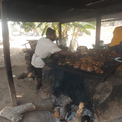 The scene depicts an outdoor food preparation area in Assai Sabongida, Nigeria, characterized by a large, active grill under a simple dark-roofed shelter. A dark-skinned male individual, wearing a dark beanie, a light t-shirt, and a green apron, is positioned centrally, actively tending to a substantial pile of roasted or grilled meat on a large metal grate. His right arm is extended towards the meat, indicating manipulation or serving. The grill structure, constructed from stacked bricks or mud, is heavily blackened with soot from prolonged use. Burning wood logs and embers are visible within the lower portion of the grill, providing heat. On the ground around the grill are several cut logs of wood. A second individual, wearing a yellow shirt, is partially visible on the far right, behind the meat pile. In the background, beyond the shelter, an unpaved area reveals several parked or moving vehicles, including large trucks and cars, and a motorcycle, suggesting a roadside or market environment. Bright daylight illuminates the scene, with trees and vegetation visible further in the background. A blue plate with food is on the far right side of the grill.