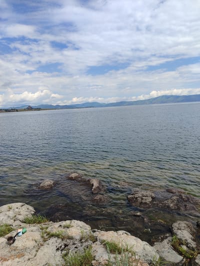 The image depicts a serene lakeside scene in Berd, Armenia. In the foreground, large, weathered rocks are interspersed with patches of green grass. Two distinct objects lie on the rocks: a small, dark metal pot and a blue and green object, possibly a cup or a container, connected by a white cord. The water's edge is visible, with submerged rocks and clear water allowing a glimpse of the lakebed. The lake itself stretches into the distance, its surface rippled by a gentle breeze. Beyond the lake, rolling green hills form a distant horizon, topped by a pale blue sky with scattered white clouds. On the far shore, a small town with buildings is nestled against the foothills, and a larger, modern-looking structure with a red roof is visible on the left side of the frame. The overall impression is one of natural beauty and tranquility.