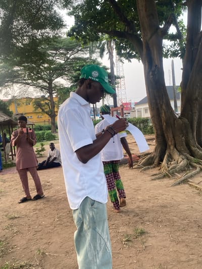 A group of people gather outdoors under a tree. One individual in the foreground wears a green NYSC cap, a white shirt, and light-wash jeans, holding a pen and paper. Another person nearby wears a white shirt and colorful patterned pants. Someone in a brown outfit stands to the side holding a phone. The background features trees, buildings, and construction equipment, suggesting an urban setting. The scene is casual, possibly a meeting or event outdoors.