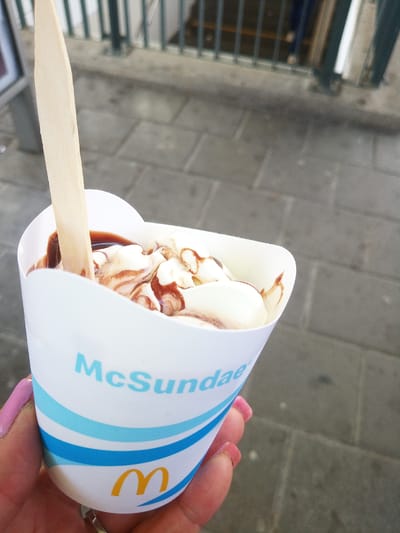 In a close-up, eye-level shot, a hand with pink fingernails holds a white paper cup containing a McSundae. The ice cream in the cup is topped with a drizzle of chocolate sauce. A light-colored wooden spoon is inserted into the ice cream. The cup has blue and white stripes and features the text "McSundae" in blue letters, along with a yellow McDonald's logo at the bottom. The background consists of gray paving stones and a dark metal railing, suggesting an outdoor urban setting, possibly in Bad Homburg vor der Höhe, Germany.