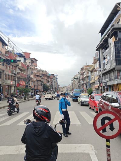 In Gokarneshwar, Nepal, a traffic policeman in a blue uniform and white gloves directs traffic on a busy street. He is positioned on a pedestrian crossing, holding his right hand up as if signaling to stop. The policeman is wearing a face mask and a police cap.
The street is lined with multi-story buildings on both sides, featuring balconies, air conditioning units, and various signage. A line of vehicles, including cars, motorcycles, and a truck, are stopped or moving slowly in traffic. A red car and a red vehicle are prominent in the foreground.
On the right side of the road, a circular traffic sign displays a black U-turn arrow crossed out with a red diagonal line, indicating no U-turns are permitted. The sky is cloudy. People are seen on the sidewalks, some sitting and others walking, as well as riding motorcycles. The scene captures a typical urban traffic management situation with ongoing vehicular and pedestrian activity.