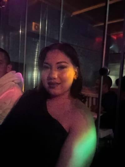 The scene depicts an indoor, dimly lit environment, identified as a nightlife establishment in the city of Dolni Tsibar, Bulgaria. A female subject is positioned in the foreground, looking towards the camera with a slight smile. She has dark hair and is wearing a dark, possibly off-the-shoulder garment. Her face is illuminated by a yellowish light, while her neck and chest display pinkish-red tones, and her right arm/shoulder exhibits a green light. To the far left, a partial view of a male subject's head and shoulder is visible, clad in a light-colored, textured garment. The background consists of multiple vertical reflective panels, creating fragmented views. Visible within these reflections or in the background are an orange square fixture in the upper left, horizontal wooden slats in the mid-right, and a second male figure, seated and partially obscured, wearing a dark shirt. The overall lighting scheme is artificial and multi-colored, predominantly purple, red, and green.