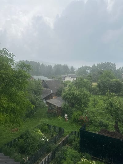 This media file captures an outdoor scene with a focus on the natural environment and some human-made structures. The overall impression is that of a somewhat overcast day with precipitation, likely rain or perhaps even snow, given the white specks visible throughout the image.
The scene is dominated by lush green trees and foliage, suggesting a warm season, possibly summer or late spring. In the background, several houses with grey, tiled roofs are visible nestled amongst the trees, indicating a rural or semi-rural setting. A dark, angular structure, possibly a fence or a shed, is present in the right portion of the image, with a section of it appearing dark and reflective. There are hints of a garden or cultivated area with some flowering plants, including white blossoms and red flowers, adding pops of color to the greenery. A thin, light-colored line, perhaps a string or wire, is visible extending across the middle ground.
The white specks scattered across the image are the most prominent detail, suggesting falling precipitation. This event creates a slightly hazy or diffused atmosphere, obscuring distant details and contributing to a sense of being immersed in the weather. The lighting is soft, consistent with an overcast sky, and there is no strong indication of a specific time of day beyond it being daytime.
The location context provided is Postoyata, Ukraine, which helps to situate the scene within a specific geographical region. There is no visible text within the image. The overall mood is one of quiet observation of nature's elements and the surrounding environment, with a subtle feeling of coziness or domesticity due to the presence of the houses.