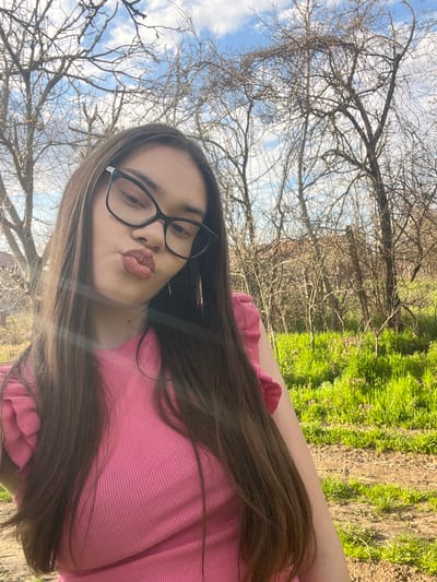 A woman with long brown hair, wearing glasses and a pink top, poses for a selfie. She is puckering her lips. The background shows bare tree branches against a partly cloudy sky and a patch of green grass. The location is likely outdoors in a park or wooded area, however, no landmarks are identifiable. The exact location in Blaskovo, Bulgaria cannot be determined from the image.