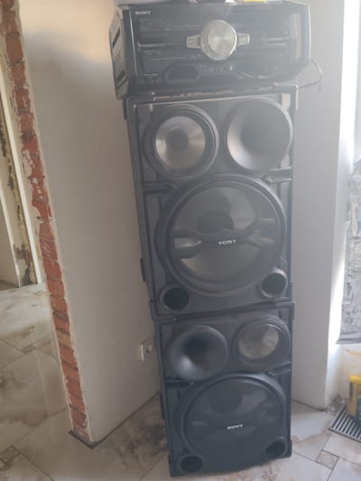 A tall, black Sony stereo system is set against a pale, textured wall. The stereo unit is composed of multiple stacked sections. The topmost section appears to be a CD player and receiver with a prominent silver dial and various buttons. Below this are several speaker units. The largest speaker, positioned in the middle, has a circular speaker grille with the "SONY" logo visible in white lettering. There are other smaller circular speaker drivers and what appear to be bass reflex ports on the other speaker sections. The overall impression is of a powerful audio system.
The scene is indoors, likely a room undergoing renovation. To the left of the stereo, a brick wall is exposed, showing unfinished plaster and insulation. The floor is tiled with a beige and brown patterned design. The lighting suggests it is daytime, with natural light illuminating the right side of the stereo system, creating some glare and bright spots. A white electrical outlet is visible on the wall behind the stereo. To the right of the stereo, some objects on the floor are partially visible, including what might be cleaning supplies or tools, and a striped mat. The setting appears to be a domestic or commercial space in the process of being worked on. No people are visible in the image.