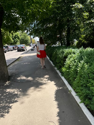 A woman with long brown hair walks away from the camera down a concrete sidewalk. She is wearing a white t-shirt and a red skirt. She has a small white purse slung over her shoulder. She is walking on the right side of a street in Sarny, Ukraine. The street is lined with green bushes and trees, with cars parked and driving on the road. Shadows from the trees are cast on the sidewalk.