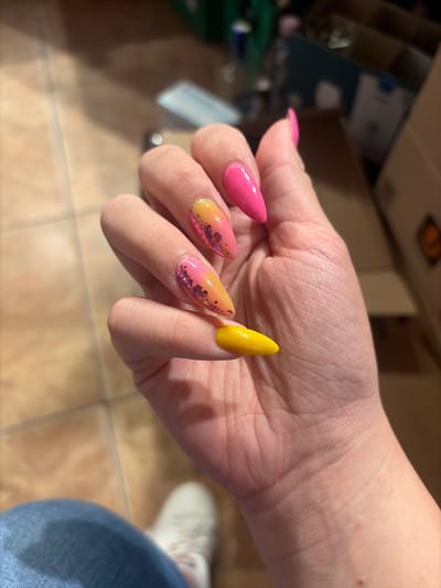 A close-up shot focuses on a person's hand with brightly colored, almond-shaped acrylic nails. The nails are painted in a gradient of pink and yellow, with pink glitter accents on the ring and middle fingers. The thumb nail is a solid bright pink, while the index finger is a vibrant yellow. The background is slightly out of focus, showing what appears to be an indoor setting with tiled flooring and the suggestion of cardboard boxes and some personal belongings. The lighting suggests it could be daytime, with sunlight casting shadows. The scene is casual and focuses on the aesthetic of the nails. No visible text is present. The location is identified as Beguntsi, Bulgaria.