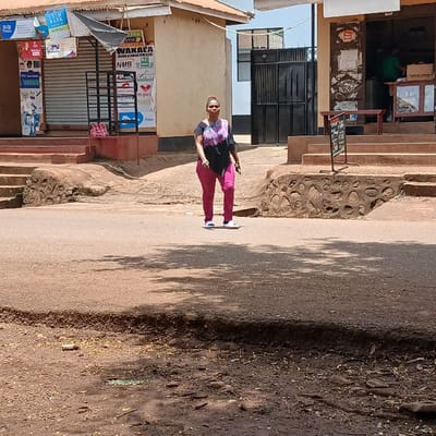 A sunlit street scene in Mji Mpya, Tanzania, features an adult woman walking on an asphalt road. The woman, of medium build, wears a black short-sleeved top with a purple/pink pattern, bright pink trousers, and white slip-on sandals, moving towards the viewer's left in the mid-ground. The immediate foreground is unpaved reddish-brown dirt with scattered debris. On the left, an elevated light-brown building with a reddish sloped roof displays multiple commercial signs, including "EXIM BANK WAKALA," "NMB WAKALA," and "m-pesa." One storefront is secured by a metal roller shutter, and a red-and-white plaid bag rests on the building's reddish steps. A low stone retaining wall with embedded circular stones borders this elevated area. On the right, an adjacent elevated building with a darker facade and a similar reddish sloped roof presents an open shop entrance. Inside, a person wearing a green shirt is visible behind a counter. A black sandwich board sign stands near the building's reddish steps. In the background, a darker structure with a large black metal double gate is visible under a clear blue sky. Tree shadows are cast on the road surface.