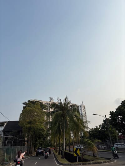 This image depicts a street scene in Binong, Indonesia, with a focus on a road and its surroundings under a bright, hazy sky, suggesting it's daytime, possibly late morning or early afternoon. The weather appears overcast or polluted, creating a muted, diffused light.

The scene features several key subjects:

*   **Vehicles:** A dark-colored SUV is prominent on the road, followed by several motorcycles. One motorcycle, in the foreground on the right, carries two people. The rider appears to be pointing off-frame, perhaps at something of interest or in communication with someone. Another motorcycle further up the road has a rider in a green jacket and helmet, suggesting they might be a delivery rider.
*   **People:** Two people are visible on the motorcycle in the foreground, one driving and the other as a passenger. The passenger is wearing a light-colored jacket and has long dark hair.
*   **Vegetation:** Lush green trees and several palm trees are scattered throughout the scene, framing the road and buildings. The palm trees, with their distinctive fronds, are particularly noticeable.
*   **Buildings:** A multi-story white building with many windows stands behind the palm trees, and another, taller building is partially visible in the upper right. A dark-tiled roof of a building is also visible in the lower part of the image.
*   **Street Furniture and Cues:** A street lamp is visible on the left side of the road. A yellow and black chevron sign is placed on the side of the road, indicating a directional warning or hazard.

The overall activity seems to be typical urban traffic movement. The presence of motorcycles suggests a common mode of transportation in the area. The image captures a moment of daily life with a focus on the urban environment, its infrastructure, and its inhabitants going about their routines. No specific emotions are overtly conveyed, but the scene is active with the flow of traffic. The license plate on the motorcycle in the foreground reads "B 6640 JSH".
