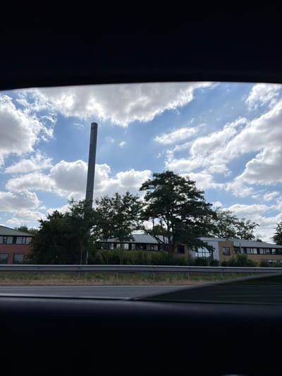 The image, taken from inside a vehicle, captures a view of a modern building complex with a distinctive tall, slender smokestack or ventilation tower in the foreground. The building appears to be a school or office building, with a long, low-rise structure featuring numerous windows. Mature trees are abundant around the complex, some partially obscuring the facade of the building. A metal guardrail separates the building area from what appears to be a road or highway in the foreground. The sky above is a bright blue with fluffy white clouds, suggesting a clear, possibly sunny day. The perspective suggests the photo was taken while driving past the location. This scene is consistent with the vicinity of a large institution or industrial area within a suburban or semi-industrial part of Évry-Courcouronnes, France, possibly near a transport artery.