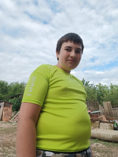 A young person, appearing to be of Caucasian ethnicity, stands outdoors and looks directly at the camera. They are wearing a bright neon green short-sleeved t-shirt with "TOUR" printed vertically in white on the left sleeve. Their hair is dark brown and cut short. The background suggests a somewhat unkempt rural or semi-rural environment, possibly on the outskirts of Leușeni, Moldova. There are trees and some discarded materials, including what looks like wooden structures, concrete blocks, and metal debris. The sky is partly cloudy with a bright blue hue visible. A small bird is flying in the distance. The overall impression is of a casual, candid outdoor portrait.