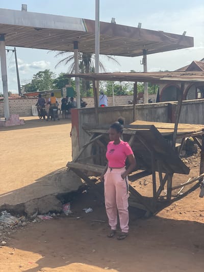 The image shows a woman in a pink shirt and light pink pants standing in an area with construction or scaffolding in Ikoga, Nigeria. She is facing left of the frame. There is dirt and trash on the ground to her right. In the background are buildings and people, along with palm trees under a sunny sky.