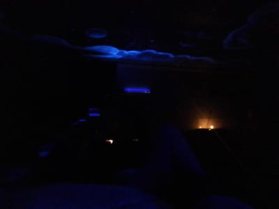 The image depicts a dimly lit indoor environment, characteristic of a relaxation room or spa, situated in Long Beach, United States. The upper portion of the scene features an overhead structure composed of irregularly shaped, billowy forms illuminated with a diffuse blue glow, resembling stylized clouds, along with several distinct circular blue light elements. In the mid-ground on the right side, multiple small, spherical light sources emit a warm orange-yellow glow from a low-lying surface. Towards the mid-ground left, several horizontal linear blue light sources are visible, receding into the background. The immediate foreground on the lower left displays a dark, indistinct form, consistent with a draped object or a reclined human leg, subtly illuminated by the ambient blue light. No other individuals or specific actions are discernible within the frame.
