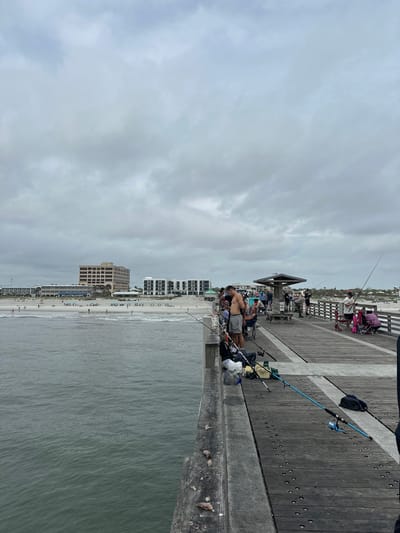 The photograph shows a scene at the Jacksonville Beach Fishing Pier in Jacksonville Beach, Florida, United States of America. The pier extends from the beach out into the ocean. The image is taken from the pier, looking towards the beach and cityscape. A group of people are fishing from the pier. The beach is crowded with people, and several tall buildings are visible along the shoreline. The sky is overcast with gray clouds. The pier itself is wooden with a gray concrete railing. Fishing rods and equipment are seen along the pier. A man is standing at the railing, apparently fishing.