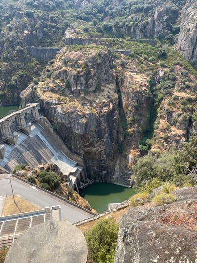 This image provides a high-angle view of the Miranda do Douro Dam (Barragem de Miranda), located within a deep, rocky gorge in Miranda do Douro, Portugal. The primary subject is a large, light grey concrete arch-gravity dam with a stepped downstream face, spanning the narrow canyon. To the left, a expansive body of green-tinted water forms the reservoir, while a smaller, similar-colored pool is visible at the dam's immediate base downstream. The dam is flanked by steep, rugged rock cliffs, primarily dark grey and brown, with sparse, dry-looking green and brown vegetation clinging to the slopes and more dense foliage in the upper and shaded areas. A paved access road extends along the dam's crest and curves downwards along the left bank. No individuals are present, and no actions or interactions are observed within the scene. The environment is characterized by arid, rocky mountainous terrain under clear daylight conditions. In the bottom-left foreground, a circular concrete or stone structure is partially visible, indicating a high vantage point for the photograph.