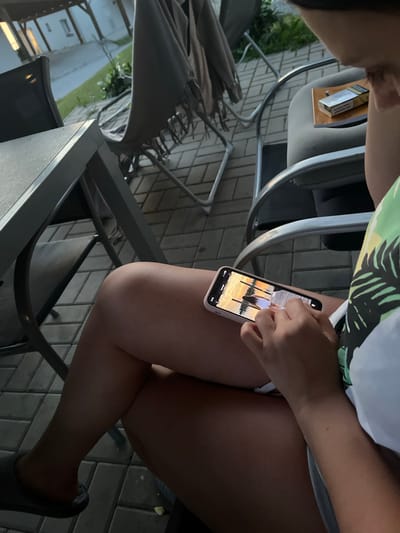 The image shows a person sitting on a chair outdoors, likely on a patio with a brick-paved floor. They are holding a smartphone in their hands, looking at the screen. On the screen, a photograph of a person standing on a wooden pier near a body of water at sunset is displayed. In the background, there are outdoor chairs and a table, suggesting a dining or lounging area. A pack of cigarettes and a lighter are placed on the chair next to the person. The lighting suggests it is late afternoon or early evening. The location is identified as Milná, Czechia.