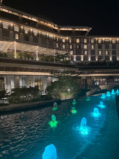 This media file presents a captivating nighttime scene of a modern architectural complex, likely located in Dehradun, India. The primary subjects are tall, contemporary buildings and a decorative water feature.

The main building, possibly a hotel or residential complex, rises prominently on the left, characterized by numerous square windows, many of which are illuminated from within, creating a warm glow. Its facade is a light color, possibly concrete or stone, accented by linear architectural lighting along its ledges and structural elements. Another building, slightly recessed, is also visible. Lush green trees and shrubs are strategically placed around the base of the buildings and along the edges of the water feature, adding natural elements to the urban setting.

In the foreground, a long, dark water body, possibly a decorative pool or canal, runs parallel to the buildings. The surface of the water is disturbed, creating ripples and reflections. Floating or submerged within this water are several striking light installations. Some of these objects glow a vivid blue, resembling abstract jellyfish or glowing organic forms, while others emit a vibrant green light. These colorful lights illuminate the surrounding water, creating dynamic patterns and reflections.

The scene is set at night, evidenced by the very dark sky and the reliance on artificial lighting to define the structures and landscape. The architectural lighting on the buildings, combined with the colorful installations in the water, creates a sophisticated and serene ambiance. No people are visible, and no specific activity is taking place. There is no visible text in the image. The overall impression is one of modern design and tranquil illumination.