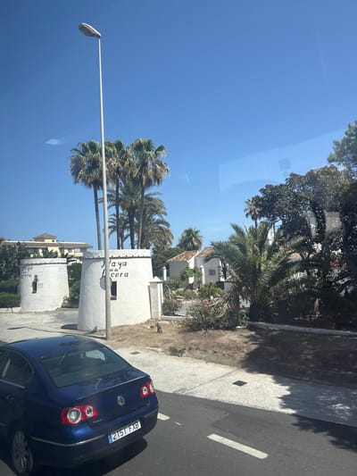 The image captures a sunlit scene in Mijas, Spain, featuring two white, cylindrical structures resembling windmills, each with an inscription that appears to read "Playa de la Cera". These structures are situated in a coastal area, with palm trees and lush vegetation surrounding them. In the background, a multi-story building with a light-colored facade is visible. In the foreground, a dark blue sedan is parked on a paved road. The sky is clear and blue, indicating a bright, sunny day. The overall ambiance is one of a tranquil, Mediterranean setting.