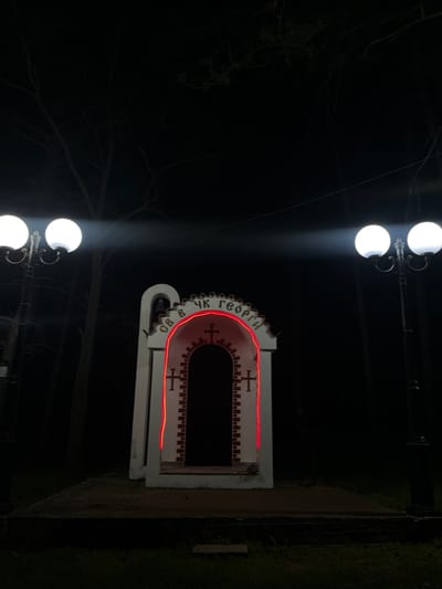 The image depicts a religious monument at night in Sungurlare, Bulgaria, under the illumination of two double-globe street lamps. The monument, outlined in red light, features Cyrillic script on its rounded top and crosses on its facade. It has a narrow, dark opening. The surrounding environment is dark with trees visible in the background.