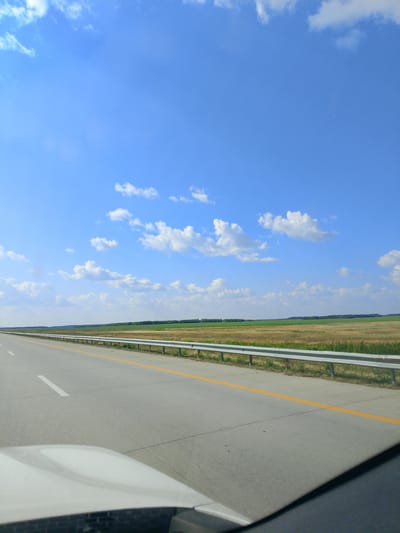 This is a photograph taken from inside a vehicle, looking forward and slightly to the right, while traveling on a highway. The highway is paved and has clear lane markings, including a white line on the left and a solid yellow line to the right of the lane. A metal guardrail runs along the right side of the highway. Beyond the guardrail, there is a flat landscape with fields of dry grass and green vegetation, possibly crops or trees, stretching to the horizon. The sky is a bright blue with scattered white cumulus clouds. The lower part of the image shows the hood of the vehicle, which is white. The scene appears to be in a rural area in Kazakhstan, possibly in the Қараөзек ауылдық округі, given the open landscape and typical highway infrastructure seen in such regions. There are no people or other vehicles visible in the immediate frame.