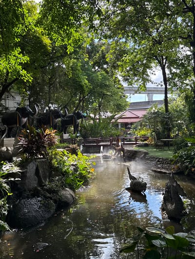 The scene depicts a garden in Samrong Nuea Subdistrict Municipality, Thailand, with a pond as a central feature. The pond contains sculptures of birds and what appears to be warm water, suggesting hot springs. Trees surround the area, creating a lush environment with natural rocks and plants. A bridge crosses the pond, and a traditional Thai-style house is visible in the background. In the distance, an elevated highway is seen above the treeline. There are decorative elephant sculptures near the ground level.