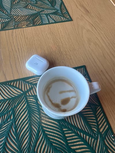 The scene is an overhead view of a wooden table surface located in Giebułtów, Poland. Two dark green placemats with an intricate leaf-cutout design are laid on the table. In the lower right portion of the frame, a white ceramic mug containing a light brown beverage, consistent with coffee and milk, exhibits frothy patterns on its surface. To the left of the mug, a white charging case, likely for wireless earbuds, is positioned on the wooden table. The initials "JB" are visibly embossed on the lid of this case. No individuals, direct actions, or interactions are depicted.