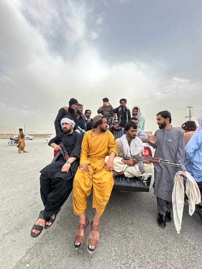 A group of men are riding in the back of a truck on a paved road in Spezand, Pakistan. One man in black clothing is holding an AK-47 rifle. Other men wear traditional attire, including loose tunics and sandals. A man on a motorcycle is visible in the background. The sky is overcast.