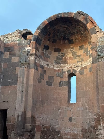 The image shows a detail of the ruins of a medieval Armenian church, specifically the Church of St. Gregory of Abughamrents in Ani, a ruined medieval Armenian city now located in present-day Turkey. The photograph is taken from a low angle, looking upwards at the structure.
The focal point is the cylindrical dome of the church, constructed from alternating light and dark-colored stone blocks. The dome's exterior features a row of decorative elements. The interior of the dome is visible, showing the concentric rings of stone that form its structure.
To the right of the dome, a section of the church wall remains, also constructed from light and dark stone. A rectangular window is visible in the wall.
The sky is visible through the window and partially surrounding the structure, indicating the building is in ruins. The overall impression is one of aged construction.