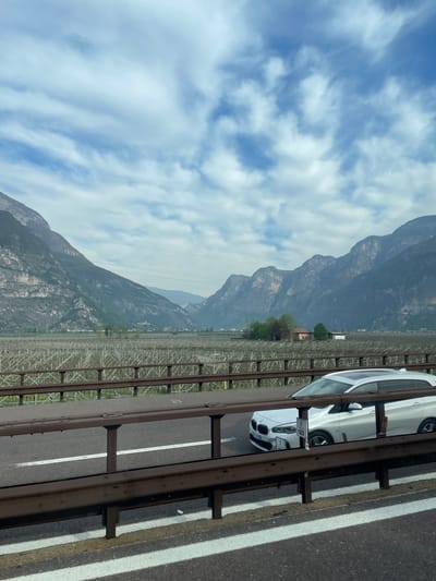 The image depicts a view from a bridge in Nave San Rocco, Italy. In the foreground, a white BMW car is seen parked on the bridge's shoulder, separated from the traffic lane by a metal barrier. The highway asphalt shows white lane markings. In the background, a mountainous landscape under a partly cloudy sky dominates the view. A large agricultural field with trellises is also visible, suggesting a vineyard or orchard. Power lines stretch across the scene.