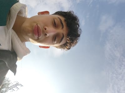 A close-up, low-angle portrait features an individual with medium complexion and dark, curly hair positioned in the upper left of the frame. The subject is wearing a light-colored hooded garment over a dark outer layer. The eyes are partially closed, and the lips are slightly parted in a pout. Direct sunlight illuminates the right side of the subject's face. The background consists of a clear blue sky with scattered white clouds. In the lower-left background, a dark, silhouetted utility pole structure is visible, along with minimal dark, out-of-focus foliage. This scene is located in Mankat al-Hatab, Syria.
