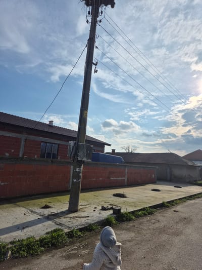 A low-angle shot captures a sunny outdoor scene in Mechkarevo, Bulgaria. In the foreground, an indistinct figure of a child stands along the edge of a concrete curb and weathered asphalt road. Behind them, a red brick building extends toward the right, with a visible window and a blue object on the roof. The building's exterior is adjacent to the concrete curb, which is interspersed with patches of green grass. An aged utility pole is positioned prominently in the frame, extending diagonally from the lower left to the upper center. Multiple wires extend from the top of the pole, bisecting the view of a partly cloudy sky. The clouds are white against a blue backdrop, with the sun slightly obscured. The scene is bathed in sunlight.