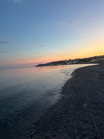 The image depicts a beach at dusk in Limenaria, Greece. The sky transitions from a clear blue at the top to pastel shades of pink and orange near the horizon. The sea is calm, reflecting the soft colors of the sky. The shoreline is covered in dark pebbles. In the background, along a hillside, are buildings and lights from the town of Limenaria. A plastic bottle is visible on the pebbled beach in the lower right portion of the image. There are no people visible in the scene.
