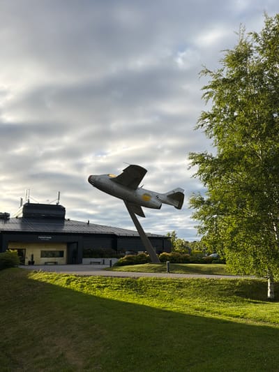 A vintage Swedish fighter jet, a Saab 29 Tunnan, is mounted on a pole in front of the Frösö Park Hotel & Spa in Östersund, Sweden. The jet is depicted mid-flight, angled upwards with its nose pointing towards the sky. The hotel building is dark and modern, with a sign indicating "FRÖSÖ PARK HOTEL & SPA". A lush green lawn slopes in the foreground, bathed in sunlight with a distinct shadow stretching across it. To the right, a vibrant green birch tree stands tall, its branches reaching towards a dramatic cloudy sky. The image captures a serene outdoor setting with a blend of historical aviation and contemporary architecture.