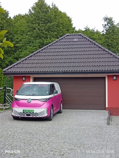 In the city of Wilczyce, Poland, on June 9, 2026, at 12:53 PM, a bright pink and white Volkswagen ID. Buzz van, bearing the license plate DW-8WU41, is parked in the driveway of a red garage with a brown sectional door. The van has "FUSSY WAGON" written on its windshield. The driveway is paved with gray interlocking stones, and the surrounding environment is dominated by lush green trees and a dark gray tiled roof on the garage. The scene appears to be a residential area, with no people visible.