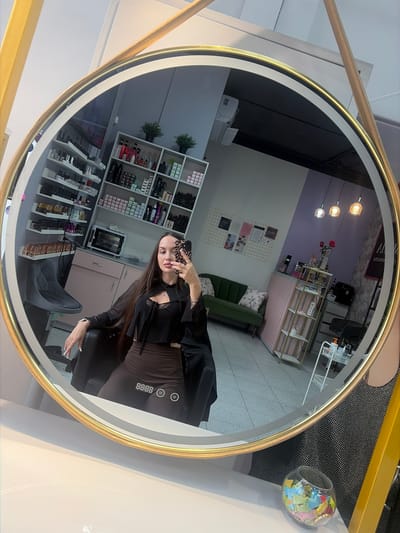 A full-body, reflected selfie of a woman in a salon is captured in a circular mirror with a gold frame. The woman, with long dark hair, is wearing a black cutout top and brown shorts. She is seated in a black chair, holding a phone in her right hand to take the picture. The salon's interior is visible in the mirror's reflection, showing shelves stocked with beauty products, a green sofa, a pink reception desk, and hanging lights. The background includes tiled floors and walls. The scene appears to be a beauty salon.
