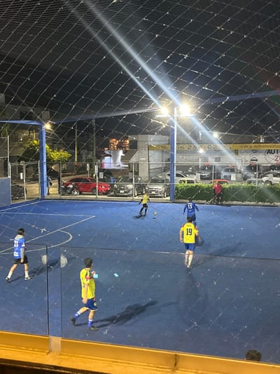 An overhead shot reveals a futsal game in progress on a blue court enclosed by netting. Three players in blue and yellow jerseys are visible, with one dribbling the ball. A fourth player in blue is positioned further down the court. Exterior buildings and parked cars are visible behind the netting on one side of the court. Bright stadium lights illuminate the playing area.