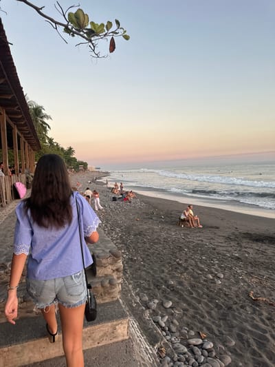A woman stands on steps overlooking a black sand beach at sunset. People relax on the beach and at the water's edge. Gentle waves approach the shore. The woman wears a blue striped shirt and denim shorts. A black purse hangs over her shoulder. Palm trees and a rustic structure are visible on the left. The sky fades from blue to orange near the horizon.