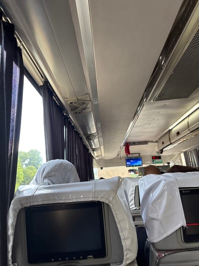 The image shows the interior of a bus, looking forward from a passenger's seat. Dark curtains are drawn on the left side, revealing a view of green trees outside. In the foreground, the back of a white seat cover with a tablet screen is visible. Another similar seat cover is visible to the right. The ceiling of the bus has overhead compartments and air vents. A small screen is mounted on the ceiling further ahead, displaying a blue interface. The overall scene suggests a journey on a bus, possibly in a tropical or temperate climate given the foliage. The location in Santa Cruz, Philippines, cannot be determined from the visual information alone.