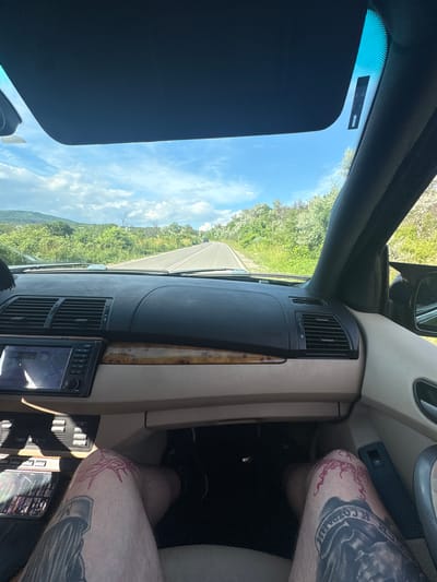The image is taken from the interior of a car, looking forward through the windshield at a paved road winding through a rural, green landscape under a partly cloudy sky. The dashboard of the car is visible, with an infotainment screen displaying a map. The front passenger's legs are visible in the lower part of the frame, showing tattoos on the knees and shins. The context provided states the location is Golemo Buchino, Bulgaria.