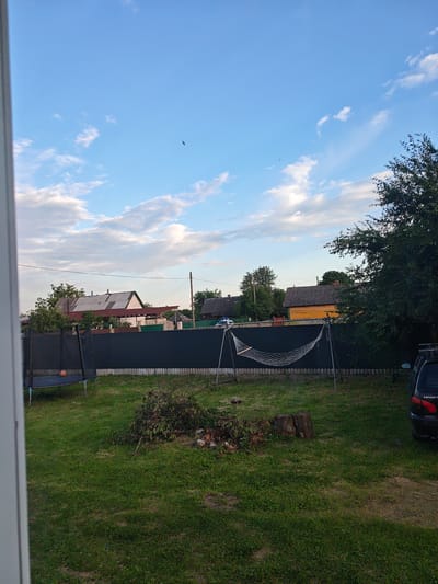 The image depicts a backyard scene in a rural area, likely in or near Гарочыцкі сельскі Савет, Belarus. In the foreground, a patch of green grass is visible, with some scattered branches and two tree stumps. A trampoline with an orange ball on it stands to the left, and a hammock is strung between two poles in the middle ground. Behind the trampoline and hammock, a dark, horizontal fence stretches across the scene. Beyond the fence, several houses with various roof types are visible, along with trees and utility poles. The sky is blue with scattered white clouds, and a single bird is flying in the distance. A dark-colored car is parked on the right side of the image. The overall impression is one of a quiet, domestic setting.