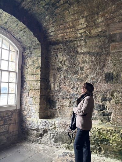 An adult female stands in profile, facing right, within a historic stone chamber, likely Blackness Castle, located in Blackness, United Kingdom. She wears a light brown tweed-patterned jacket with a dark, possibly faux-fur, collar, and dark trousers, holding a dark shoulder bag in front of her. Her head is tilted upwards, and her gaze is directed towards the arched stone ceiling and the textured, rough-hewn stone wall. The walls and ceiling are constructed from large, irregular blocks of grey and brown stone, exhibiting areas of green moss or dampness, indicative of age and environmental exposure. A large, arched window with multiple white-framed panes dominates the left side of the frame, providing significant natural light that illuminates the chamber and the subject. To the right of the window, a rectangular stone recess is built into the wall. The floor is composed of large, irregular grey stone flags. The subject is observing the upper architecture or details of the stone structure.