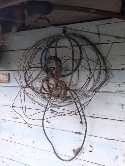 An outdoor scene in Deremezna, Ukraine, features a weathered wooden wall with peeling white paint. A tangled mass of various wires and cables, some appearing rusty and others insulated in brown and blue, hangs suspended from a horizontal rope attached to the wall. A small, rusted metal plate is visible on the wall to the left of the hanging wires. The overall impression is one of disuse and storage of electrical components.