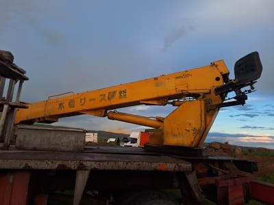 Anipemza, Armenia: An aging, yellow crane is mounted on a weathered truck bed, with some rust and possible deterioration. The crane is positioned against a backdrop of a cloudy sky tinged with dusk colors. The crane has Japanese text. In the background, there are several vehicles, including a white van with its hood open, and at least one person standing near the van, possibly performing maintenance. The surrounding terrain is dry with sparse vegetation.