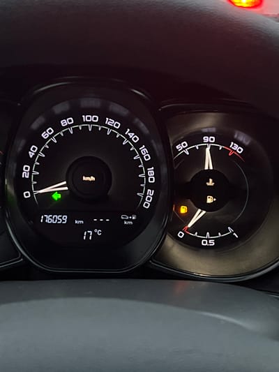 This is a close-up, vertically oriented shot of a car's dashboard, likely taken in Ижевск, Russia. The image focuses on the instrument cluster, which includes a speedometer, fuel gauge, and a digital display. The speedometer is a prominent circular dial with markings from 0 to 200 km/h. The needle is currently pointing very close to 0, indicating the car is stationary or moving very slowly. Within the speedometer's center, there are two illuminated icons: one resembling a vehicle with an arrow pointing to the side, and another that looks like a fuel pump with a droplet. Below these, a small, orange warning light is illuminated, shaped like a gas cap with a diagonal line through it, suggesting a fuel system issue or an open gas cap. To the right of the speedometer is a fuel gauge, with markings from 0 to 0.5. Its needle is also very close to the 0 mark, indicating a low fuel level. Below the speedometer, a larger digital display shows several pieces of information. The odometer reads "176059" kilometers, and next to it are units of "km". Above this, there's a temperature reading of "17°C". There are also icons and markings indicating kilometers per hour. A green arrow is pointing downwards in the lower part of the digital display. The overall scene is dark, typical of a car interior, with the illuminated dashboard being the primary light source. The steering wheel or dashboard trim is visible on the left and right sides of the image, made of dark, textured material. The faint glow of a red light is visible in the upper left corner, likely another indicator or light source within the car. The image appears to have been taken during the day or evening, but the interior lighting dominates the scene. The notable details are the low speed indicated by the speedometer, the low fuel level, the illuminated fuel system warning light, and the current outside temperature of 17°C. The odometer reading of 176,059 km provides information about the vehicle's usage. The context of Ижевск, Russia, is provided as a location cue. --- **Object/Scene:** Car dashboard (instrument cluster). **Activity:** Vehicle is stationary or moving very slowly. **Notable details:** Speedometer, fuel gauge, odometer (176059 km), temperature (17°C), fuel system warning light, illuminated icons for vehicle status and fuel. **Location Cues:** Ижевск, Russia (provided context). **Time of Day/Weather:** Interior lighting dominates, suggesting daytime or evening. **Visible