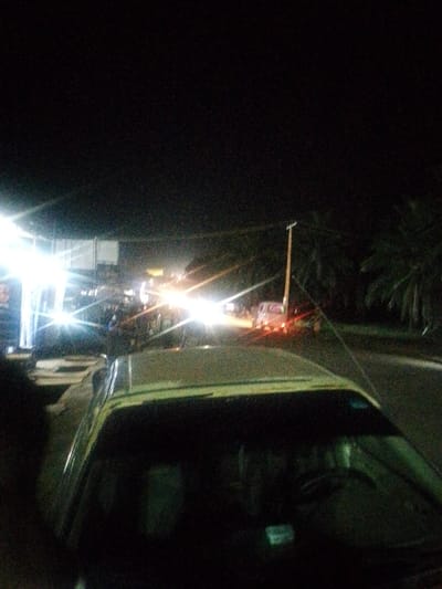 The image captures a nocturnal urban street scene in Abeokuta, Nigeria, viewed from the interior or immediate vicinity of a light-colored vehicle, likely a car, whose hood and windshield comprise the lower third of the frame. Artificial light sources heavily illuminate the midground, creating significant lens flare. To the left, a commercial establishment is brightly lit by multiple white lights, with a rectangular sign visible above. A street extends into the distance, populated by vehicles displaying bright white headlights directed towards the viewer and red taillights receding into the background, indicative of active traffic. On the right side of the street, a tall, light-colored utility pole is visible, equipped with a streetlight fixture. Dark, indistinct vegetation, resembling palm trees, borders the roadway on the right. The sky overhead is completely dark. Specific individuals are not clearly discernible, but their presence and activity are implied by the operational vehicles and commercial setting.