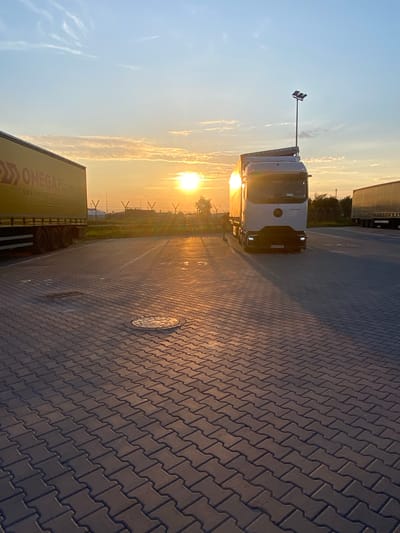 An outdoor, eye-level, medium shot captures a truck parked on a paved lot during sunset in Niepołomice, Poland. The sun, low on the horizon, casts a golden glow across the scene, illuminating the sky with soft orange and yellow hues and creating long shadows on the ground. The paved lot is made of interlocking concrete pavers, some of which appear wet, reflecting the sunlight.
In the foreground, the paver pattern is clearly visible, leading the eye towards the parked trucks. To the left, a yellow trailer with the "OMEGA LOGISTICS" logo is partially visible. In the center and slightly to the right, a white semi-truck with its headlights on is parked, facing the viewer. The truck has a Mercedes-Benz emblem on the front. A street lamp with three lights stands behind the white truck. To the right, another truck trailer is visible in the distance.
The background features a fence, vegetation, and more distant structures, all bathed in the warm, ambient light of the setting sun. The sky is a pale blue, with wispy clouds catching the sunlight. The overall impression is one of a quiet end-of-day scene at a logistics hub.