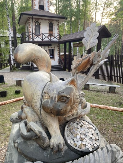 This is a full, eye-level, outdoor photograph taken in the city of Tal'tsy, Russia, featuring a large, carved wooden sculpture of a squirrel in the foreground. The squirrel sculpture is weathered and detailed, with a prominent tail arching over its back, and it is positioned on a wooden base that appears to be part of the same art installation. The squirrel's paws are positioned around a shallow wooden bowl filled with numerous coins, suggesting it might be a donation point or a wishing well.

Behind the sculpture, a rustic, two-story building with a peaked roof and white walls with brown trim is visible. This building has a balcony with wooden railings and arched windows. Adjacent to this building is a covered entryway or pavilion with a sign that is partially legible, but the word "АЗЕРБАЙДЖАН" (Azerbaijan) is discernible. The environment surrounding the sculpture and buildings consists of green grass, scattered fallen leaves, and several birch and pine trees, indicating a forested or park-like setting. Wooden benches and dark trash receptacles are also present in the background, along with a dark wooden fence. The lighting suggests it is daytime, possibly late afternoon or early evening, with some soft light filtering through the trees. No people are visible in the photograph.